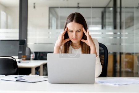 Une femme d'affaires stressée et soucieuse tient la tête dans les mains, regardant un écran d'ordinateur portable, révélant les aspects difficiles et exigeants du travail de bureau moderne. Pressions réelles du monde des entreprises
