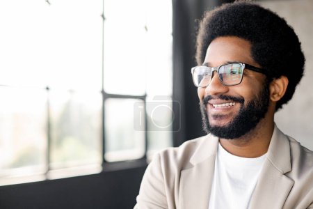 Brazilian young businessman in glasses and afro hairstyle looks aside inspired, his smart casual attire and the bright office setting portraying a vibrant, successful work culture