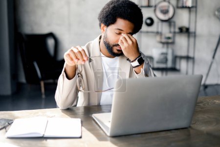 Modern businessman rubs eyes in fatigue or exasperation while working on his laptop, a depiction of the demanding nature of contemporary work life. Male office employee feels burnout and overworked
