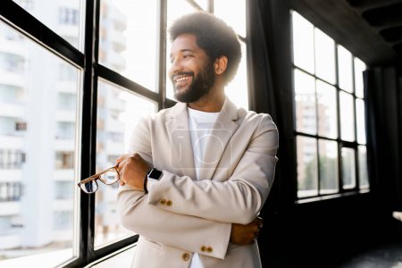 Der junge moderne Geschäftsmann steht mit einem bezaubernden Lächeln und einem Blick in die Zukunft vor einem Bürofenster und verkörpert den Geist des Unternehmertums und die Lebendigkeit der brasilianischen Unternehmenskultur