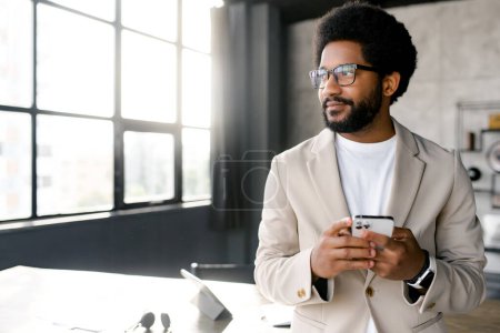 A young, stylish Brazilian businessman is standing by a window in an office setting, holding a smartphone, wearing glasses and light beige suit, looking confident and ready for a work day