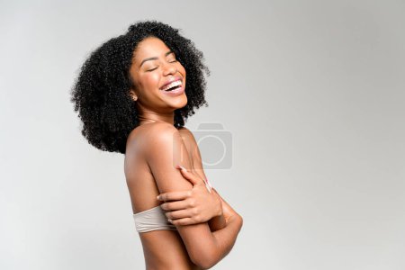 Afroamerikanerin lacht herzlich und hält den Arm über ihren Körper, ihre schwungvollen Locken umrahmen ein fröhliches Gesicht, das vor einem weichen grauen Hintergrund steht, der die unbeschwerte Stimmung unterstreicht.