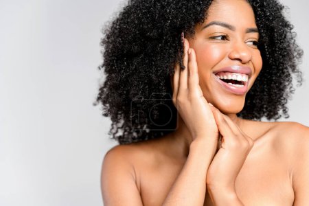 A joyful African-American woman with a vivacious smile and natural curly hair poses with her hands gently framing her face, highlighting her cheerful demeanor and radiant skin against grey background