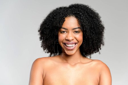 Eine überschwängliche afroamerikanische Frau mit strahlendem Lächeln und funkelnden Augen erfüllt den Rahmen vor hellgrauem Hintergrund mit purer Freude und strahlt ein offenes und ansteckendes Glück aus.
