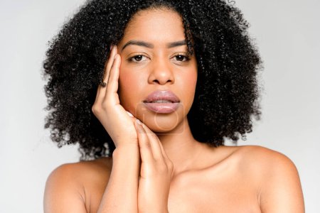 Das Nahaufnahme-Porträt einer afroamerikanischen Frau, deren Hand ihr Gesicht zart umrahmt, offenbart einen fesselnden Blick und einen Hauch reflektierter Nachdenklichkeit vor neutralem Hintergrund..