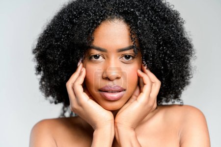 Foto de Una mujer afroamericana equilibrada con el pelo naturalmente rizado sostiene su cara suavemente, su expresión serena sobre un fondo gris suave, capturando la esencia de la belleza natural. - Imagen libre de derechos