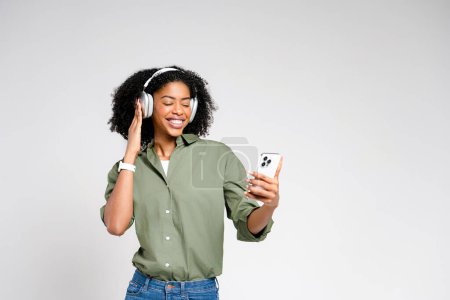 An exuberant African-American woman with a radiant smile looks at her smartphone, immersed in the world of music through her over-ear headphones, signifying a fusion of technology and lifestyle.