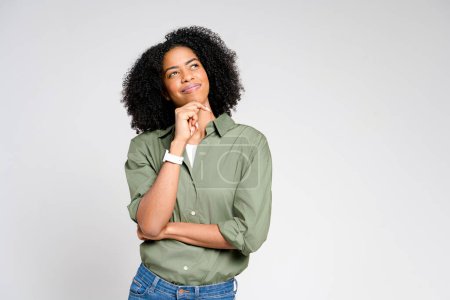 Foto de Una mujer afroamericana se para pensativamente con una mano en la barbilla, con la mirada ligeramente hacia arriba, sugiriendo contemplación o toma de decisiones sobre un fondo limpio y gris. - Imagen libre de derechos