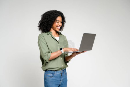 Afroamerikanerin mit warmem Lächeln arbeitet isoliert an ihrem Laptop, der die Verschmelzung von Arbeit und Komfort in einem modernen Wohnumfeld mit minimalistischer Ästhetik repräsentiert.