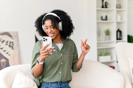 Una mujer afroamericana con una sonrisa alegre usa su teléfono inteligente mientras escucha música en sus auriculares blancos, cómodamente sentada en un sofá en una habitación elegantemente decorada.