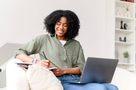 Foto de Una mujer afroamericana de contenido se involucra en múltiples tareas, escribiendo en un cuaderno mientras maneja simultáneamente las tareas en su computadora portátil en su serena oficina en el hogar - Imagen libre de derechos