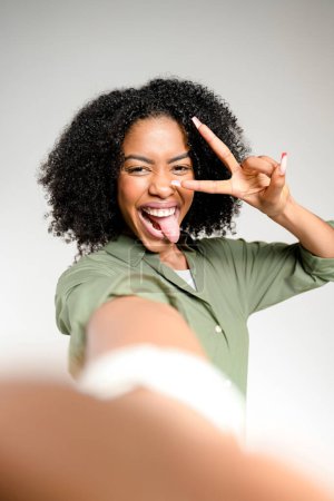Foto de Una mujer afroamericana con el pelo rizado da una señal de paz y saca su lengua en una pose juguetona de selfie, capturando un momento de diversión y espontaneidad contra un fondo gris claro. - Imagen libre de derechos