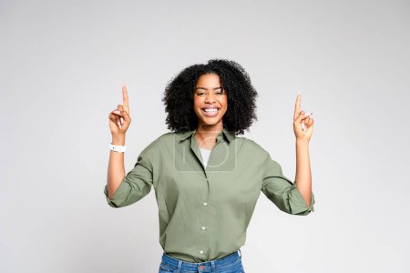 African-American woman with a beaming smile points upwards with both index fingers, exuding enthusiasm and winning attitude. Celebratory moment, perfect for conveying triumph or positive announcement