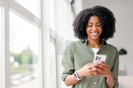 Dans un cadre intérieur bien éclairé, une femme afro-américaine au sourire contagieux interagit avec son smartphone, s'engageant avec des amis ou des followers via un réseau social.