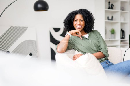 Téléchargez les photos : Une femme afro-américaine s'assoit sereinement sur un canapé, son sourire ravissant et sa tenue élégante sur fond de maison moderne et minimaliste, incarnant une atmosphère domestique chic et décontractée.. - en image libre de droit