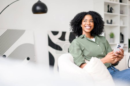Joyful African-American woman interacting with her smartphone, enjoying a pleasant online exchange, within a tastefully designed living space. The integration of technology into leisure and lifestyle