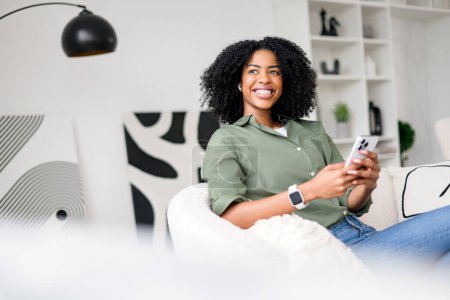 Foto de Capturando un momento de felicidad genuina, una mujer afroamericana interactúa con su teléfono inteligente, posiblemente compartiendo buenas noticias o disfrutando de una conversación humorística en una vida moderna y de buen gusto. - Imagen libre de derechos