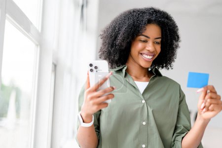 Foto de Mujer afroamericana vibra de placer mientras sostiene un teléfono inteligente y una tarjeta de crédito, posiblemente haciendo una compra o reserva en línea, en un interior brillante y moderno. - Imagen libre de derechos