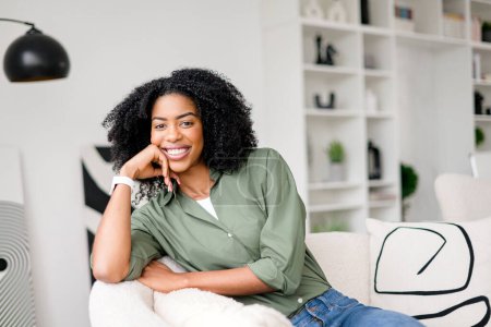 Una mujer afroamericana con una sonrisa cálida se sienta cómodamente en un sofá, su mano descansando suavemente en su cara, exudando una sensación de relajación y satisfacción en un ambiente de sala de estar brillante y moderno.