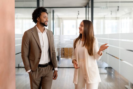 Dans un couloir de bureau moderne, un homme d'affaires afro-américain et son collègue sont engagés dans une discussion animée, marchant ensemble
