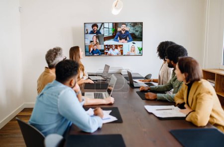 Team engaged in a virtual meeting with a display screen, showing both remote participants and in-person colleagues interacting seamlessly, integration of modern technology in collaborative workspaces