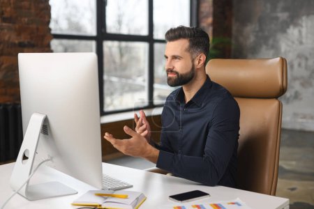 Photo for Young bearded confident successful man in business casual clothes talking during video call, skilled job applicant is ready for an online interview on a video call, sitting at the desk - Royalty Free Image