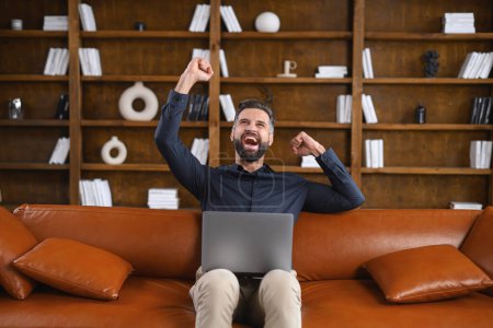 Photo for Happy excited freelancer man is sitting on a couch, smiling while using a laptop, celebrating victory or success, raisin fists in triumph gesture - Royalty Free Image