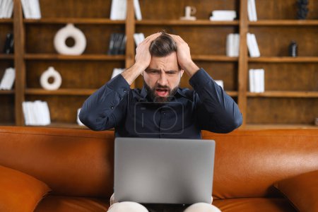 Photo for An unhappy young male freelancer looks at a laptop screen shocked by gadget breakdown or operational problems. A frustrated man confused and surprised by an unexpected error on a computer device - Royalty Free Image