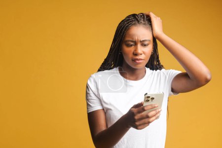 Photo for Shocked brunette young woman holding smartphone, looking at mobile phone screen and has disappointment face, received a bad news, made a mistake and a phone is locked, isolated on yellow - Royalty Free Image