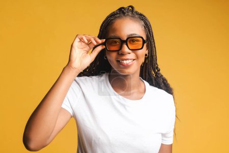 Téléchargez les photos : Jeune femme brune portant des lunettes de soleil se tient et regarde la caméra, belle femme afro-américaine positive sourit isolé sur fond jaune - en image libre de droit