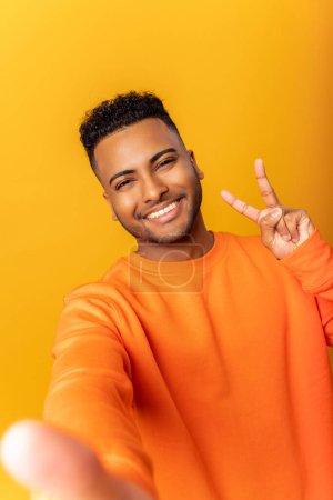 Photo for Happy satisfied man showing v sign symbol of peace with fingers, looking at camera with toothy smile, making selfie. Indoor studio shot isolated on yellow background - Royalty Free Image