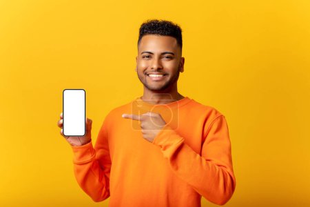 Foto de Hombre indio sorprendido sosteniendo el teléfono inteligente en la mano y señala con el dedo la pantalla del teléfono vacía, chico asombro que presenta la mejor oferta, la publicidad de la nueva aplicación móvil, aislado en amarillo - Imagen libre de derechos