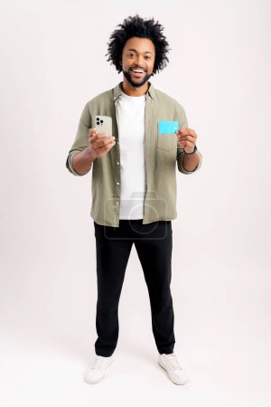 Téléchargez les photos : E-banking, transfert d'argent en ligne. homme afro-américain joyeux tenant la carte de crédit et smartphone dans ses mains isolé sur blanc, noir en utilisant l'application mobile pour le paiement en ligne, achats - en image libre de droit