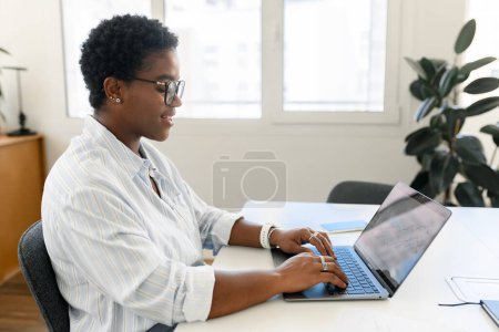 Téléchargez les photos : Indépendante afro-américaine aux cheveux courts assise au bureau avec ordinateur portable et dactylographier, écrire un article, sourire et bloguer, profiter et créer un design pour un nouveau projet, vue de côté - en image libre de droit