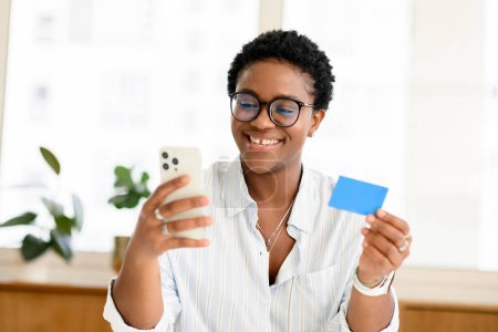 Online-Shopping. Positive junge afrikanisch-amerikanische Frau in smart lässiger Grafschaft hält Smartphone und Kreditkarte in der Hand, kauft im Internet ein, überweist online Geld