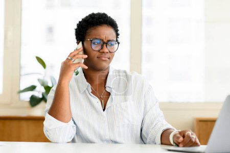 Gelangweilte afroamerikanische Büroangestellte telefoniert im modernen Büro, unglückliche schwarze Frau fühlt sich müde von der Kommunikation mit Kunden