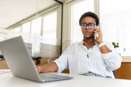 Besorgte afroamerikanische Büroangestellte, die auf dem Smartphone im Büro sitzt, Frau, die unangenehme Telefongespräche mit dem Vorgesetzten führt