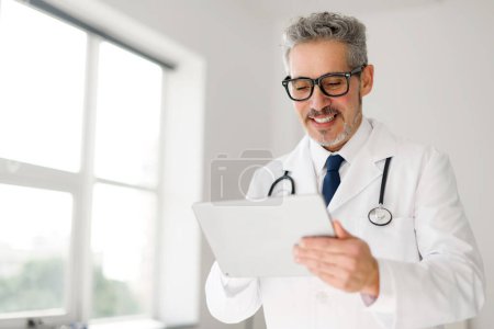 A cheerful senior doctor with grey hair and spectacles reviews information on a tablet, demonstrating the integration of technology in his daily medical practice