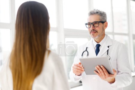 A mature doctor with grey hair discusses health information on a digital tablet with a young woman, in a clinic office filled with natural light