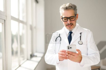 Un médico de cabello gris maduro examina su teléfono inteligente en un entorno clínico, probablemente revisando la información del paciente o las historias clínicas, destacando el uso de dispositivos móviles en la atención médica.