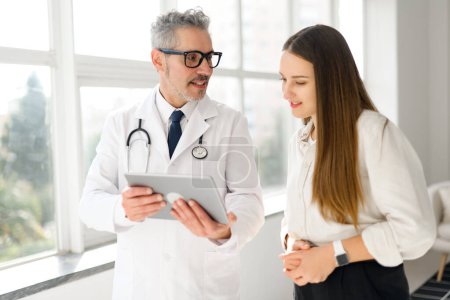 Un médico de pelo gris con una bata blanca comparte información sobre una tableta con una mujer joven en un consultorio médico moderno y brillante, encarnando un enfoque de salud colaborativo