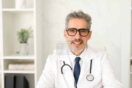 Un médecin chevronné aux cheveux gris et aux lunettes pose pour un portrait professionnel, assis dans un bureau de clinique moderne, respirant chaleur et accessibilité