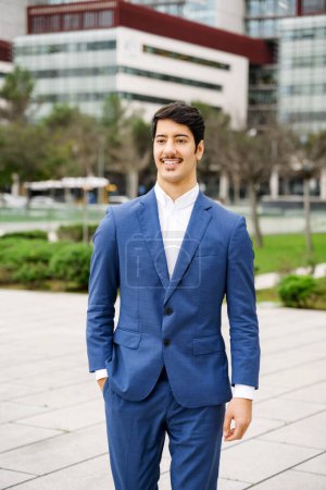 Standing with a soft smile, the Hispanic businessman enjoys a moment outside, the soft focus on the urban setting behind him illustrating a balance between professionalism and personal contentment.