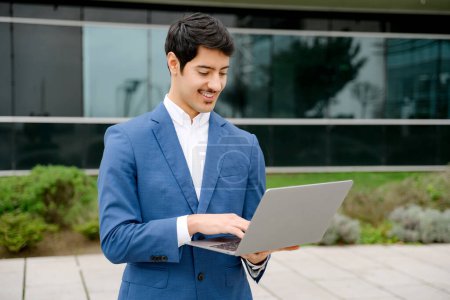 A focused Hispanic businessman in a sharp blue suit reviews data on his laptop while standing outdoors, representing the modern entrepreneur ability to work from anywhere.