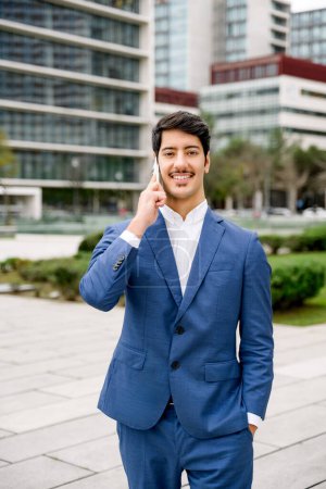 The Hispanic businessman is pictured smiling during a phone conversation, standing with confidence in a public space with the citys architectural elements in soft focus behind him.
