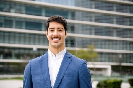 With a warm smile and a background of modern buildings, this handsome Hispanic businessman stands confidently, his blue suit symbolizing trust and professionalism.