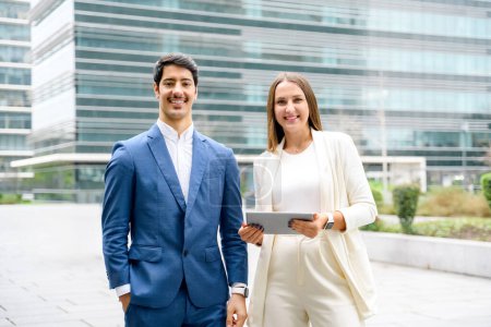 In front of glass-clad corporate building, a man and woman dressed in business attire confidently hold a tablet, looking ready to tackle their next business challenge together. Business and technology