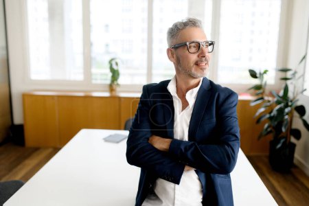Photo for A mature professional stands with arms crossed, a slight smile on his face, in a light-filled office, suggesting both experience and approachability - Royalty Free Image