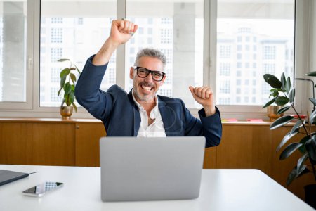 Excited mature businessman is gesturing victory with his arms raising up, happy senior ceo screaming with triumph, winning in game, having received great news of a good deal