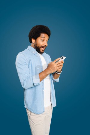 Astonished young man using his phone, expressing surprise and excitement, standing isolated on blue. This shoot captures the spontaneous reactions often elicited by digital communication.
